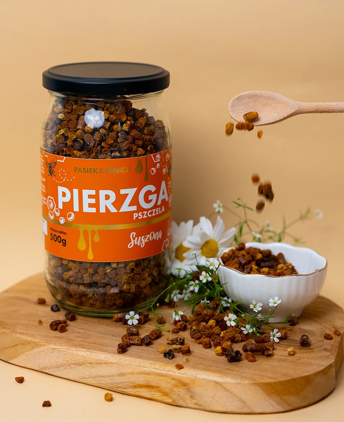 Słoik suszonej pierzgi pszczelej 500g Pasieka Mnich na drewnianej desce obok białych kwiatów.