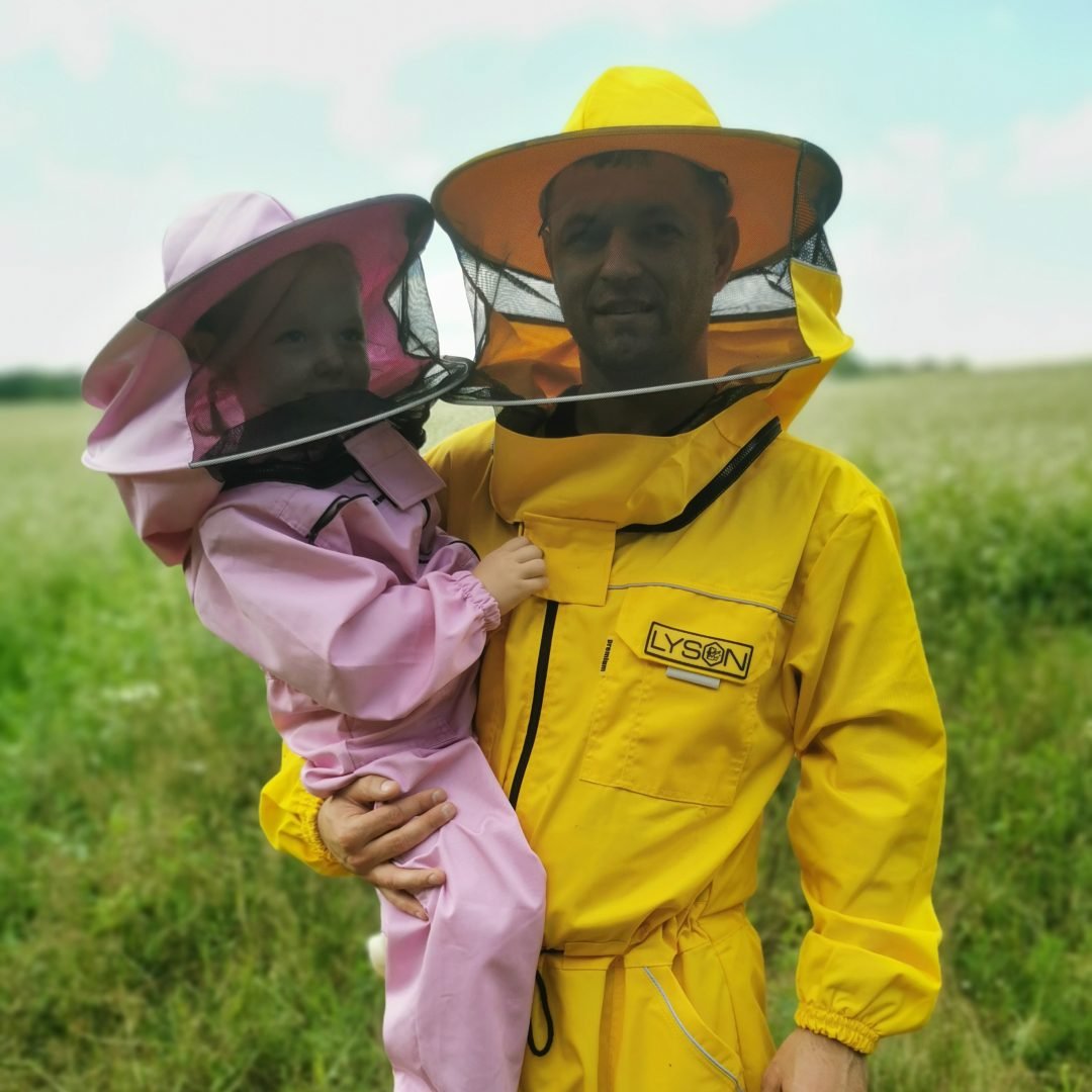 Pszczelarz w żółtym kombinezonie trzymający dziecko w różowym stroju ochronnym na tle kolorowych uli w Pasiece Mnich.